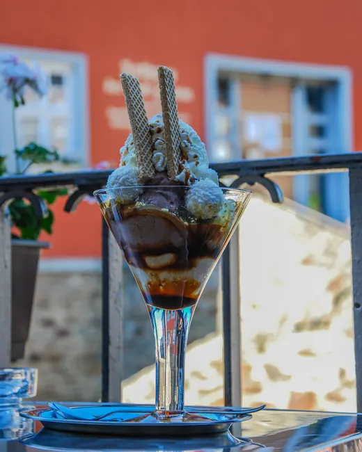Eisbecher mit Schokoladensoße und Waffelröllchen auf dem Balkongeländer, dahinter eine rote Hauswand