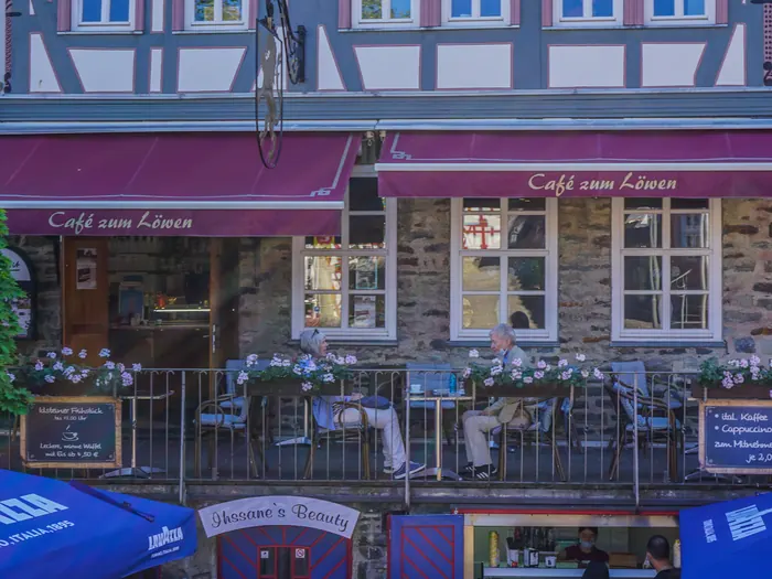 Fachwerkfassade des Café zum Löwen mit dunkelroten Markisen, Blumenkästen und Gästen auf dem Balkon