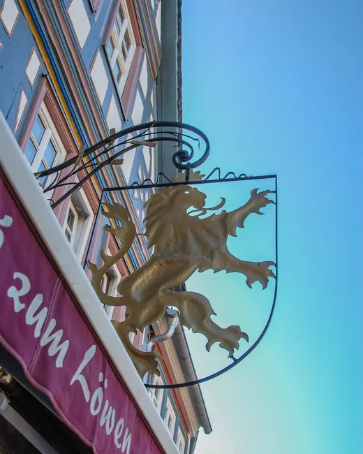 Goldener Löwe am schmiedeeisernen Wirtshausschild, darunter die Markise mit dem Schriftzug „zum Löwen“ vor blauem Himmel